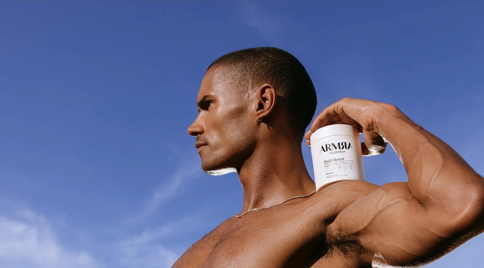Man flexing his arm with an ARMRA jar on his shoulder against a blue sky